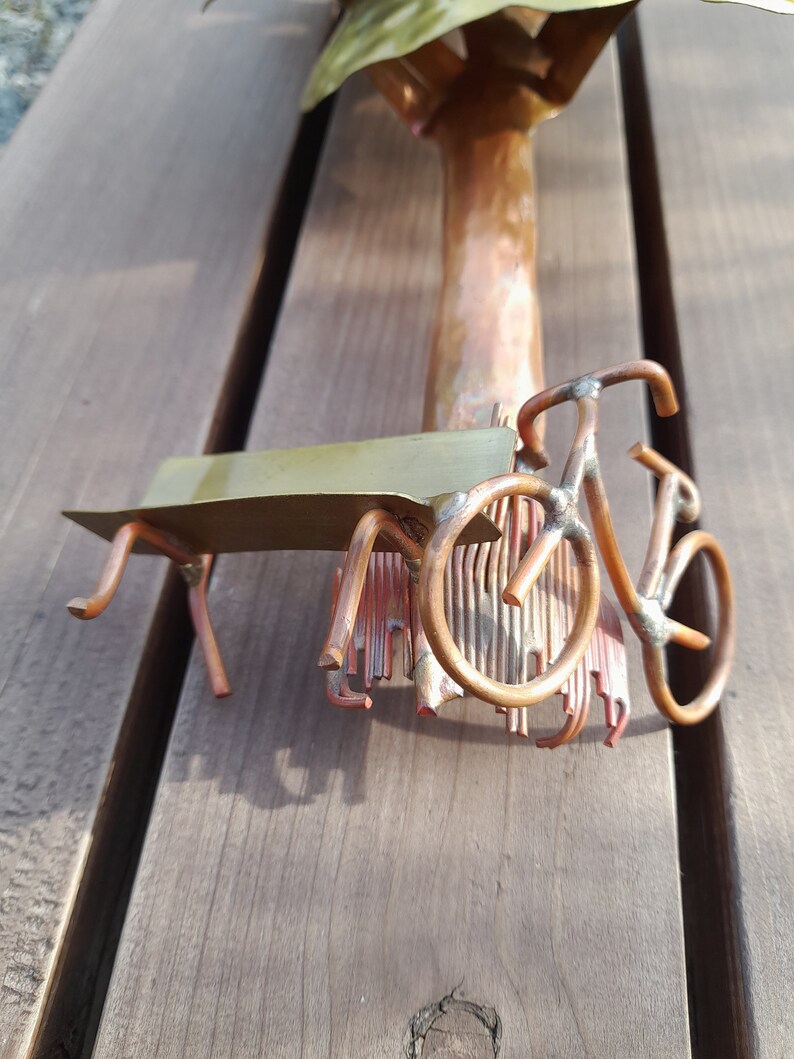 May include: A copper sculpture of a tree with a bench and a bicycle underneath. The sculpture is made of wire and has a rustic look.