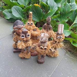 Op de afbeelding: Vijf houten beeldjes met stroomachtig haar en beschilderde gezichten. De beeldjes staan op een grijs oppervlak.