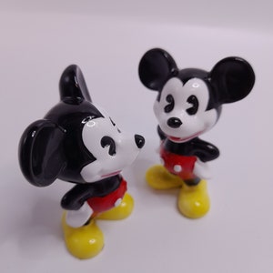 May include: Two black and white ceramic Mickey Mouse figurines with red shorts and yellow shoes. The figurines are standing on a white surface.