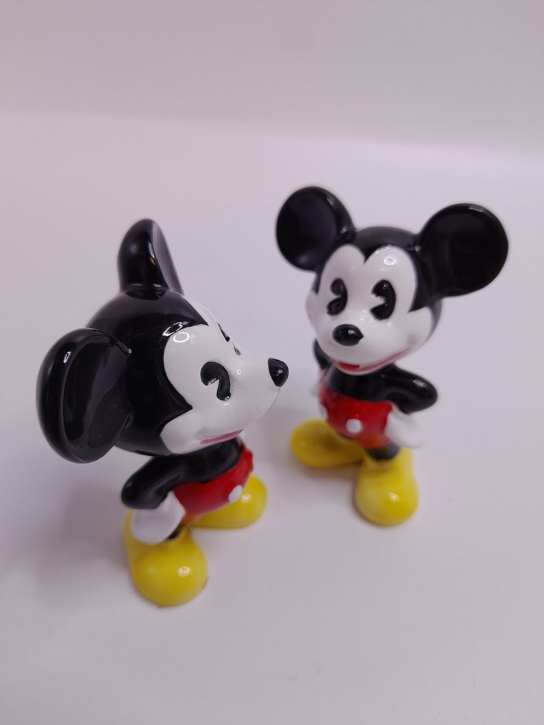 May include: Two black and white ceramic Mickey Mouse figurines with red shorts and yellow shoes. The figurines are standing on a white surface.
