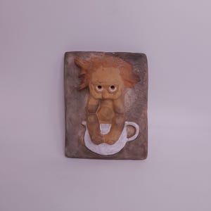 Op de afbeelding: Een grillige keramische wandplaat met een trollachtig figuur met oranje haar, zittend in een witte theekop. De figuur is lichtbruin met grote ogen en staat tegen een rechthoekige, getextureerde, grijze achtergrond.