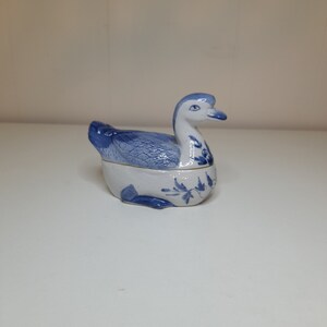 May include: A blue and white porcelain trinket box shaped like a duck. The duck has a detailed design of blue leaves and flowers.