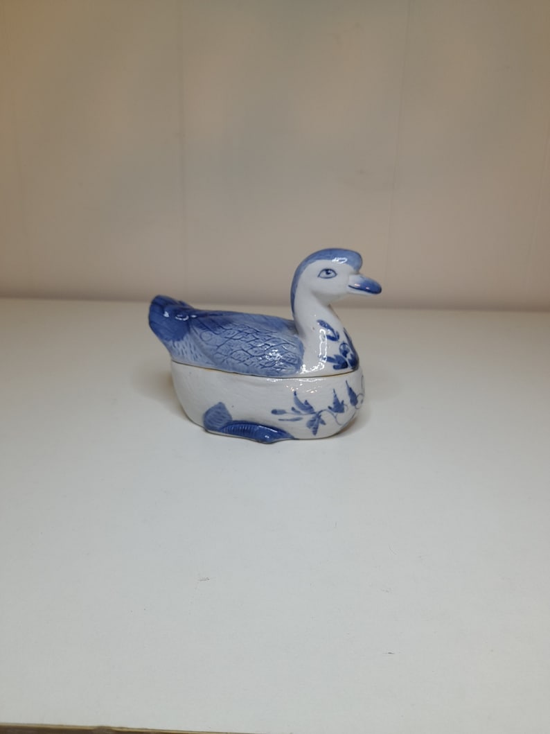 May include: A blue and white porcelain trinket box shaped like a duck. The duck has a detailed design of blue leaves and flowers.