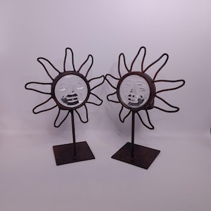 Könnte beinhalten: Zwei dekorative sonnenförmige Skulpturen. Jede hat ein Glasgesicht mit einem lächelnden Ausdruck, umgeben von einem rostigen Metall-Sonnenstrahl-Design. Sie stehen auf quadratischen Sockeln und erzeugen eine skurrile, rustikale Ästhetik.