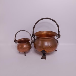 Op de afbeelding: Twee koperen ketels met gehamerde texturen en donkere metalen handvatten en poten. De grotere ketel heeft een gebogen handvat en drie poten. De kleinere ketel heeft een kleiner handvat en één poot. Beide op een witte achtergrond.