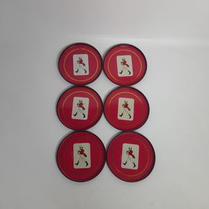 Op de afbeelding: Zes rode onderzetters met een gouden rand. Elke onderzetter heeft een witte rechthoek met een figuur in een rode jas en zwarte laarzen. De onderzetters zijn gerangschikt in twee rijen van drie.