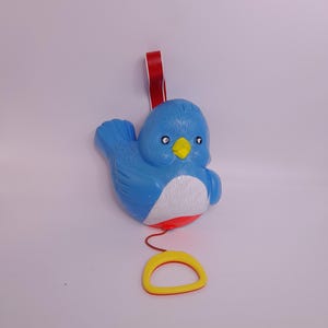 Könnte beinhalten: Ein blaues Plastikvogelspielzeug mit rotem Band und gelbem Zugring. Der Vogel hat einen weißen Bauch, einen gelben Schnabel und schwarze Augen. Das Spielzeug ist für Kinder konzipiert.