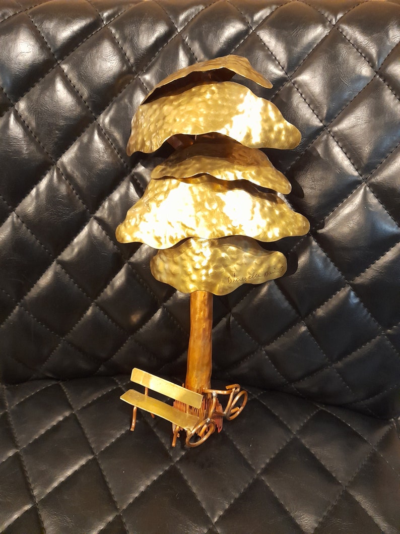 May include: A gold metal sculpture of a tree with a bench and a bicycle underneath. The tree has a textured surface and the sculpture is signed "Bruce Lu 2013".