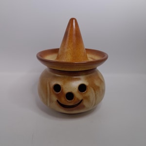 Puede incluir: Un tarro de cerámica con tapa en forma de sombrero. El tarro es de color marrón claro con un diseño de cara sonriente, con dos agujeros redondos para los ojos y una boca curvada. La tapa tiene forma de cono, también marrón claro, y se asienta sobre un ala ancha.