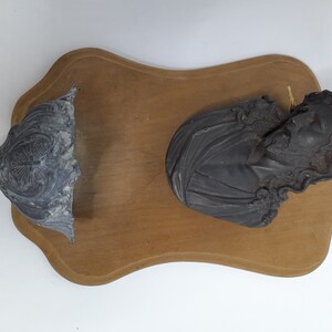 K&ouml;nnte beinhalten: Eine Holztafel mit zwei daran befestigten schwarzen Gipsfiguren. Eine Figur ist eine B&uuml;ste einer Person mit langen Haaren, die andere ist ein dekoratives St&uuml;ck.