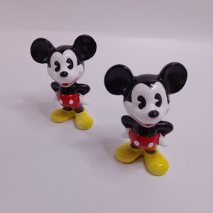 May include: Two black and white ceramic Mickey Mouse figurines with red shorts and yellow shoes. The figurines are standing on a white surface.