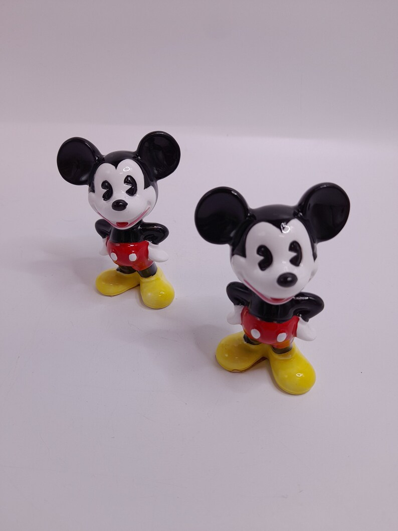 May include: Two black and white ceramic Mickey Mouse figurines with red shorts and yellow shoes. The figurines are standing on a white surface.