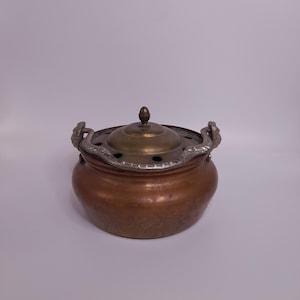 May include: A copper-colored pot with a lid and decorative handles. The lid is a brass color with a small knob on top. The rim of the pot has a silver-colored decorative trim. The pot has a round shape.