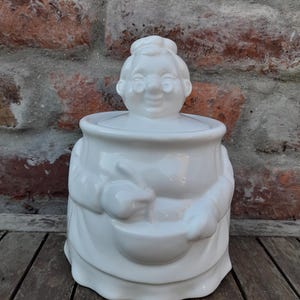 Op de afbeelding: Een witte keramieken koekjespot in de vorm van een persoon met een schort en een koksmuts. Het figuur heeft een glimlachend gezicht, een bril en houdt een mengkom vast. De pot heeft een afneembaar deksel dat het hoofd van het figuur vormt.
