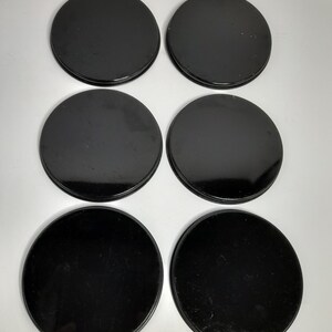 Op de afbeelding: Zes zwarte, ronde onderzetters, gerangschikt in twee rijen van drie. De onderzetters hebben een glanzende afwerking en lijken van een massief materiaal te zijn gemaakt. De onderzetters liggen op een wit oppervlak.