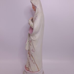 Puede incluir: Una estatua de porcelana blanca de la Virgen Mar&iacute;a, vestida con una t&uacute;nica blanca con detalles florales rosas. La estatua est&aacute; de pie sobre una base blanca con detalles florales rosas.