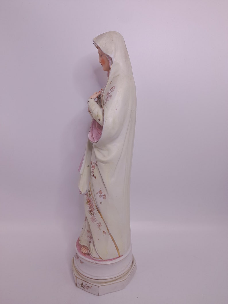 Puede incluir: Una estatua de porcelana blanca de la Virgen Mar&iacute;a, vestida con una t&uacute;nica blanca con detalles florales rosas. La estatua est&aacute; de pie sobre una base blanca con detalles florales rosas.