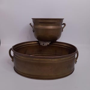 Könnte beinhalten: Zwei antike Pflanzgefäße aus Messing. Das obere Pflanzgefäß ist ein kleiner, runder Topf mit zwei Griffen. Das untere Pflanzgefäß ist eine große, oval geformte Schale mit zwei Griffen. Beide Pflanzgefäße haben ein angelaufenes, gealtertes Aussehen.