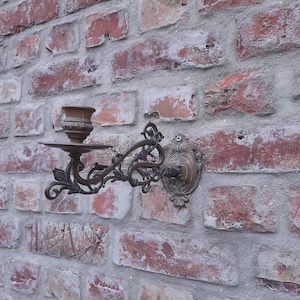 Può includere: Un'antica applique da parete in bronzo con un portacandele e decorazioni a volute. L'applique è montata su un muro di mattoni usurati, mettendo in risalto il suo design vintage. Il portacandele è progettato per una singola candela.