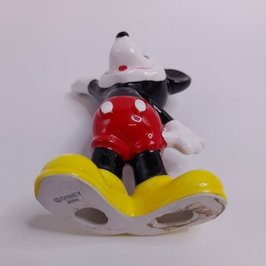 May include: A ceramic figurine of Mickey Mouse standing with his arms outstretched. He is wearing his signature red shorts with white polka dots, black shoes, and white gloves. The figurine is marked "&copy;DISNEY JAPAN" on the bottom of one foot.