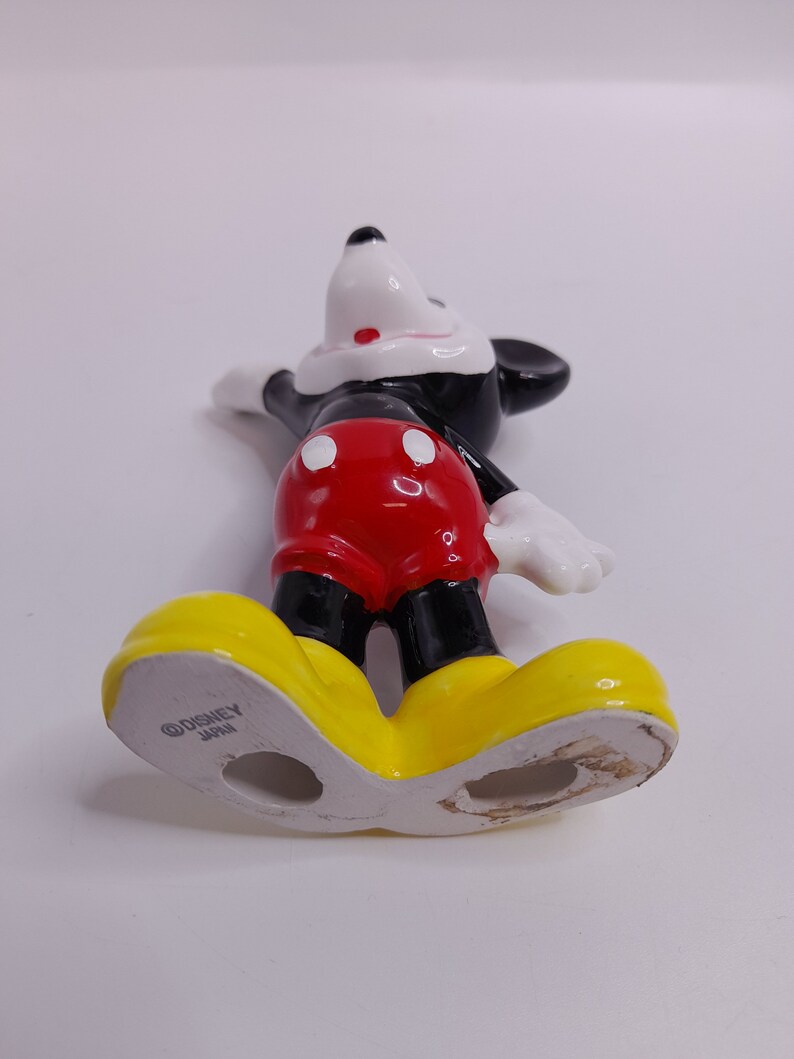 May include: A ceramic figurine of Mickey Mouse standing with his arms outstretched. He is wearing his signature red shorts with white polka dots, black shoes, and white gloves. The figurine is marked "&copy;DISNEY JAPAN" on the bottom of one foot.