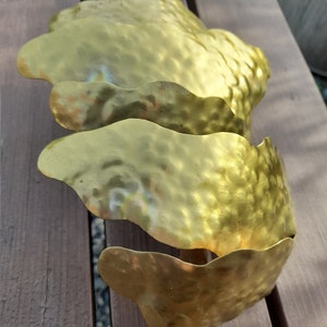 May include: Three gold metal leaves with a hammered texture, attached to a wooden stick. The leaves are arranged in a cascading pattern.