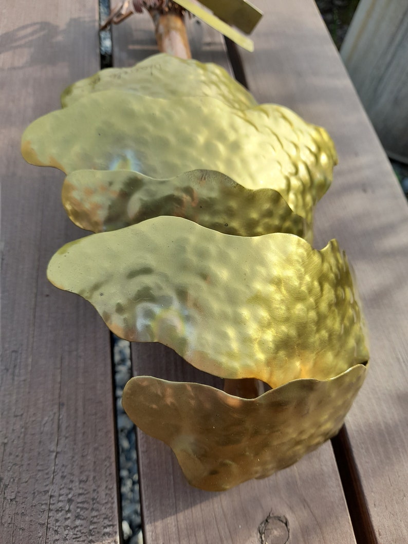May include: Three gold metal leaves with a hammered texture, attached to a wooden stick. The leaves are arranged in a cascading pattern.