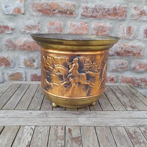 Puede incluir: Una maceta de color cobre con un relieve detallado de figuras a caballo y otras escenas. La maceta tiene un borde ancho de color dorado y se apoya en tres pequeños pies dorados. El fondo es una pared de ladrillo envejecido y una mesa de madera.