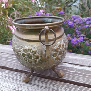 Pu&ograve; includere: Un vaso vintage in ottone con corpo rotondo, tre piedini e un manico. Il vaso presenta disegni floreali in rilievo e una patina scura. Lo sfondo comprende fiori viola e vegetazione.