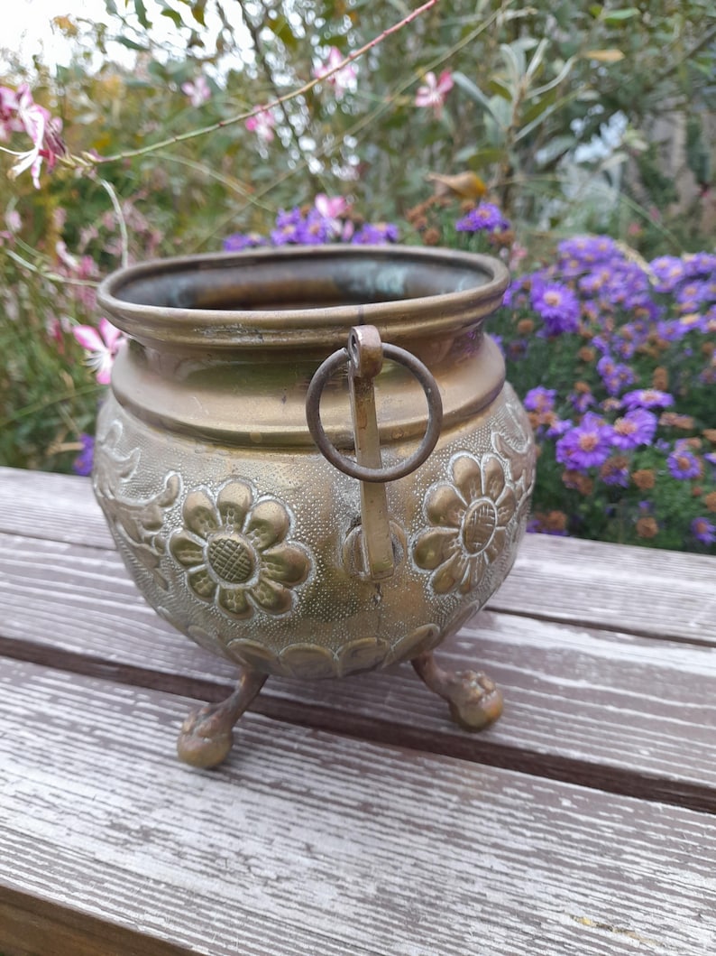 Pu&ograve; includere: Un vaso vintage in ottone con corpo rotondo, tre piedini e un manico. Il vaso presenta disegni floreali in rilievo e una patina scura. Lo sfondo comprende fiori viola e vegetazione.