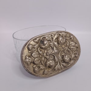 Könnte beinhalten: Ein ovaler, silberfarbener Deckel mit einem komplizierten floralen Design. Der Deckel ist aus Metall und hat ein erhabenes, geprägtes Muster.