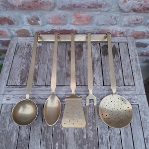May include: A set of five brass kitchen utensils hanging on a brass bar. The utensils include a ladle, spoon, slotted spatula, fork, and a slotted spoon. The utensils are displayed against a weathered wooden surface and a brick wall.