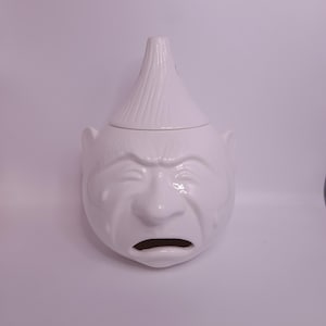 May include: A white ceramic onion-shaped jar with a lid. The jar features a sculpted face with closed eyes, a prominent nose, and an open mouth. The lid is shaped like the top of an onion with vertical ridges, and the surface is smooth and glossy.
