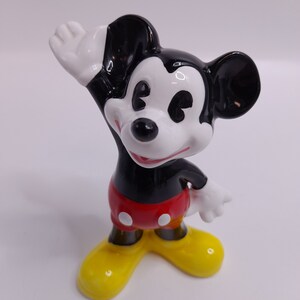 May include: A ceramic figurine of Mickey Mouse waving with his right hand. He is wearing his signature red shorts with white polka dots, black shoes, and white gloves. His ears are black and his face is white with black eyes and a black nose.