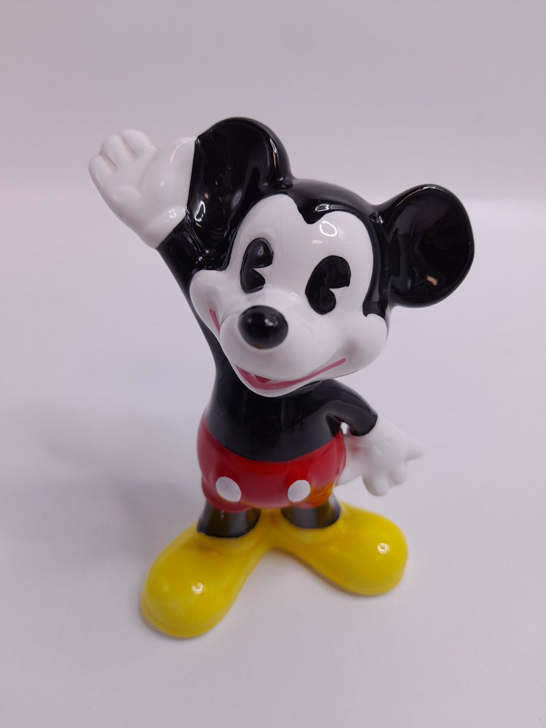 May include: A ceramic figurine of Mickey Mouse waving with his right hand. He is wearing his signature red shorts with white polka dots, black shoes, and white gloves. His ears are black and his face is white with black eyes and a black nose.