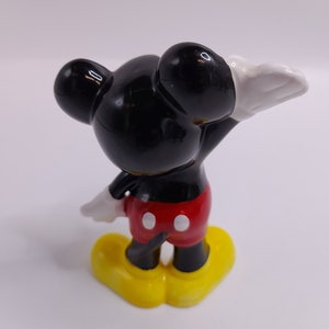 May include: A ceramic figurine of Mickey Mouse. Mickey is standing with his right arm raised and his left arm bent at the elbow. He is wearing his signature red shorts with white buttons and yellow shoes. His head and ears are black, and his gloves are white.