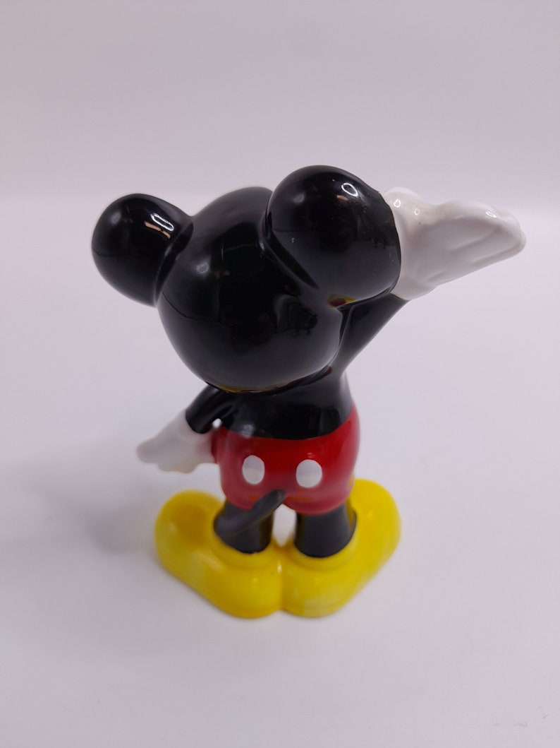May include: A ceramic figurine of Mickey Mouse. Mickey is standing with his right arm raised and his left arm bent at the elbow. He is wearing his signature red shorts with white buttons and yellow shoes. His head and ears are black, and his gloves are white.