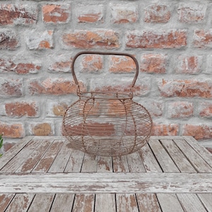 Op de afbeelding: Een antieke, roestige draadmand met een handvat. De mand heeft een ovale vorm en staat op een verweerde houten tafel. De achtergrond is een bakstenen muur.
