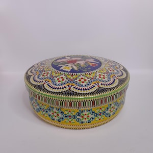 Può includere: Una scatola rotonda e decorativa con un motivo floreale sul coperchio. Il coperchio presenta un cerchio blu centrale con un'illustrazione floreale. I lati sono decorati con motivi a mosaico colorati in blu, giallo e verde.