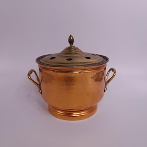Könnte beinhalten: Ein kupferfarbener Behälter mit Deckel und zwei Griffen. Der Deckel hat mehrere kleine Löcher und einen dekorativen Knopf oben. Der Behälter hat eine gehämmerten Textur und einen runden Boden. Die Griffe sind goldfarben.