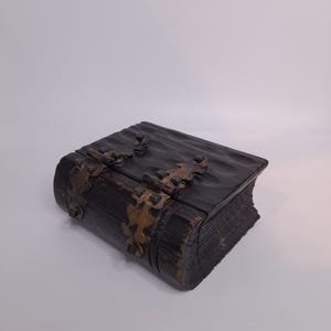 Op de afbeelding: Een donkerbruine houten doos in de vorm van een gesloten boek met een getextureerd, golvend oppervlak. De doos is voorzien van twee decoratieve, sierlijke messingkleurige metalen scharnieren aan de voorzijde die het deksel vastzetten.