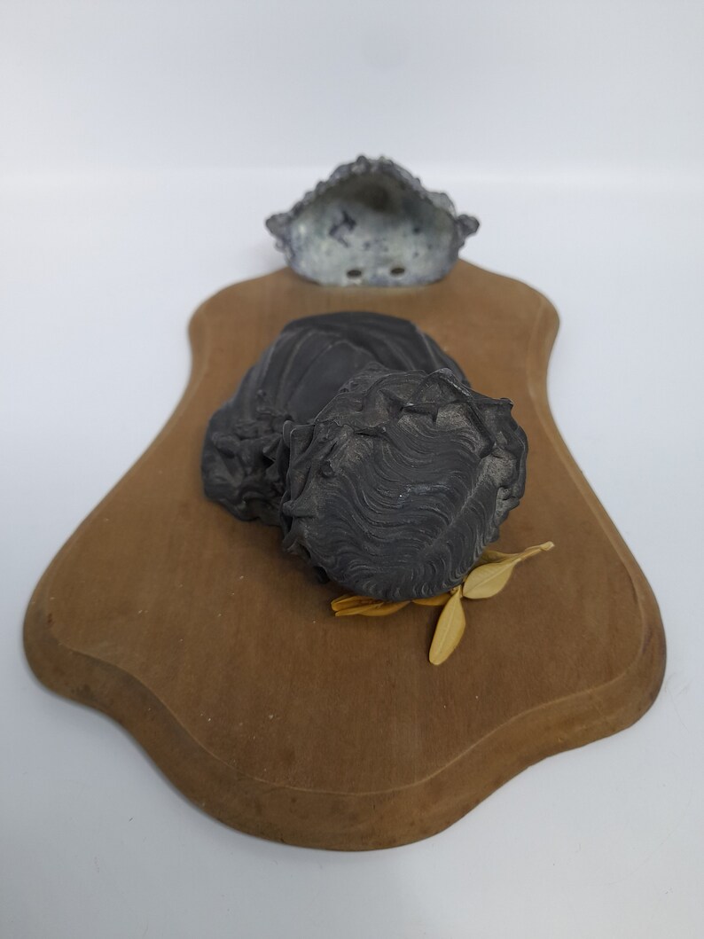 K&ouml;nnte beinhalten: Eine schwarze und graue Keramikskulptur eines Frauenkopfes auf einer Holzplatte. Die Skulptur ist auf der Platte montiert und hat eine abgenutzte Oberfl&auml;che. Die Platte ist braun und hat einen gewellten Rand.