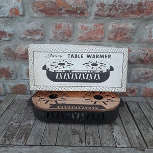 Peut inclure: Un chauffe-plat de style vintage avec une base en métal noir et doré et un dessus décoratif. Le dessus porte l'inscription "Fancy Table Warmer" et un dessin du chauffe-plat. Le fond est un mur de briques vieilli et une table en bois.