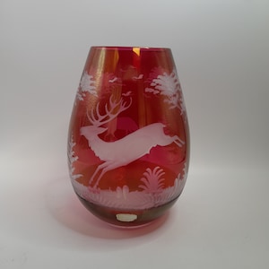 May include: A red glass vase with a white etched design of a deer running through a forest. The vase has a rounded, teardrop shape.