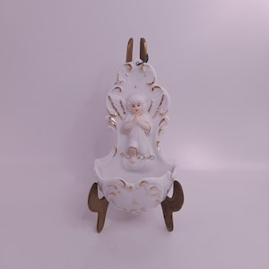 Pode incluir: Uma pia de água benta de porcelana branca com acabamentos dourados e uma pequena figura de anjo. A pia é montada em um suporte de madeira. O anjo é retratado com as mãos juntas em oração. A pia tem um design ornamentado.