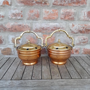May include: Two copper-colored metal containers with lids and handles. The containers have a round shape with decorative patterns and a gold-colored handle. The containers are sitting on a wooden surface.