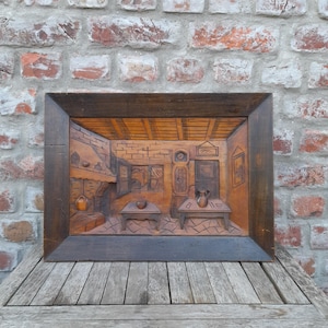 Op de afbeelding: Een rechthoekige houten wandplaat met een gedetailleerd reliëf van een rustiek interieur. Het houtsnijwerk toont een kamer met een open haard, twee tafels, een staande klok en een raam, uitgevoerd in warme bruine houttinten.