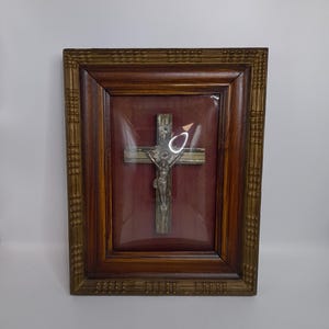 Könnte beinhalten: Ein gerahmtes religiöses Kruzifix. Das Kreuz besteht aus silberfarbenem Metall und zeigt eine Jesusfigur. Der Rahmen besteht aus dunklem Holz mit einem dekorativen Rand. Der Hintergrund ist tiefrot.
