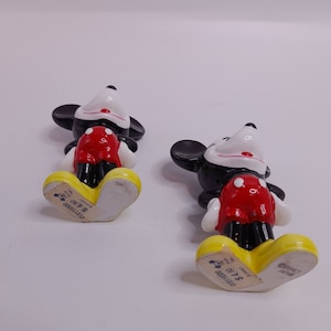 May include: Two ceramic Mickey Mouse figurines, one black and one red, standing on a white surface. The bottom of each figurine has a yellow base with the text "Disney" and "$4.99" printed on it.