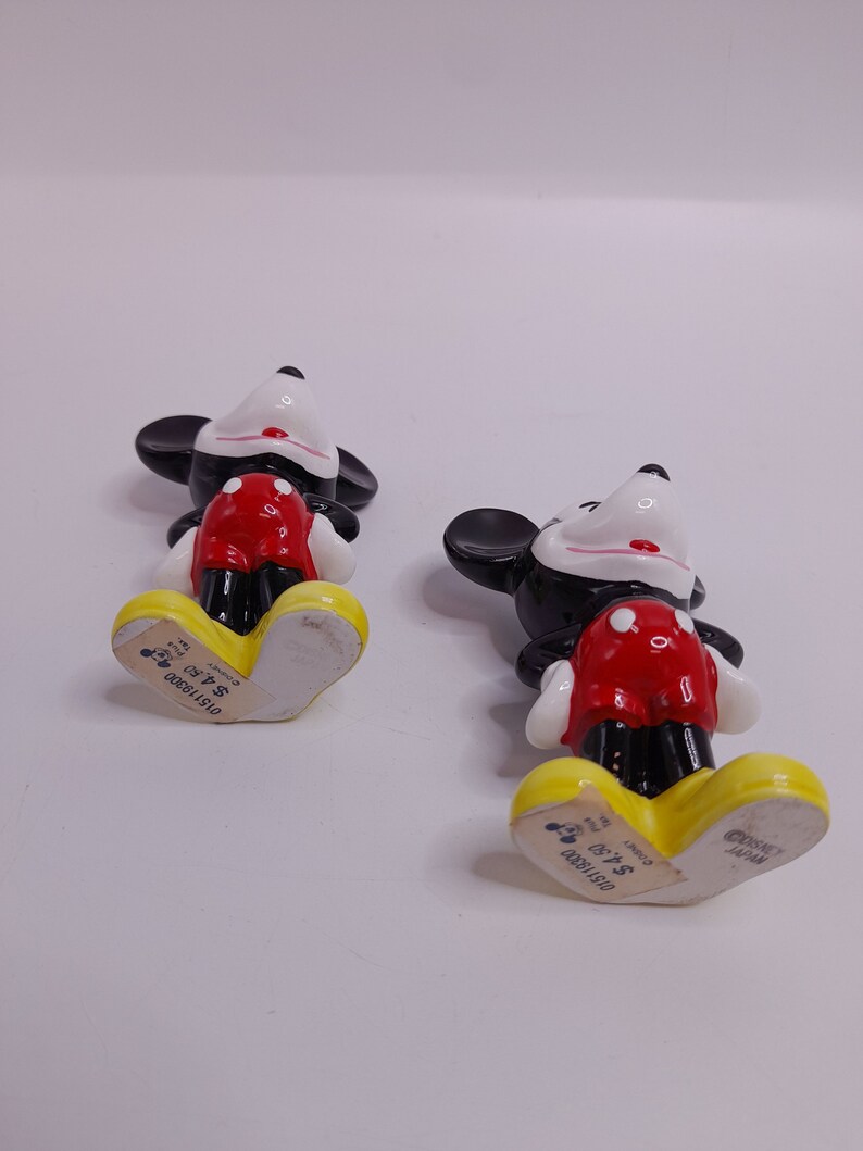 May include: Two ceramic Mickey Mouse figurines, one black and one red, standing on a white surface. The bottom of each figurine has a yellow base with the text "Disney" and "$4.99" printed on it.